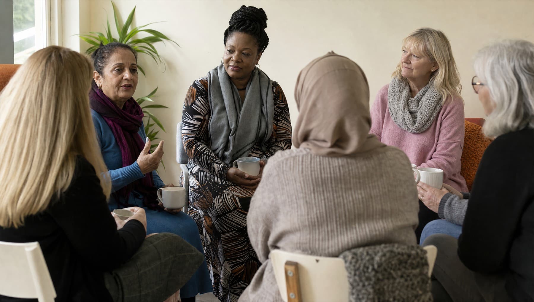 A women's peer support group sitting in a circle with mugs of tea in a bright community room